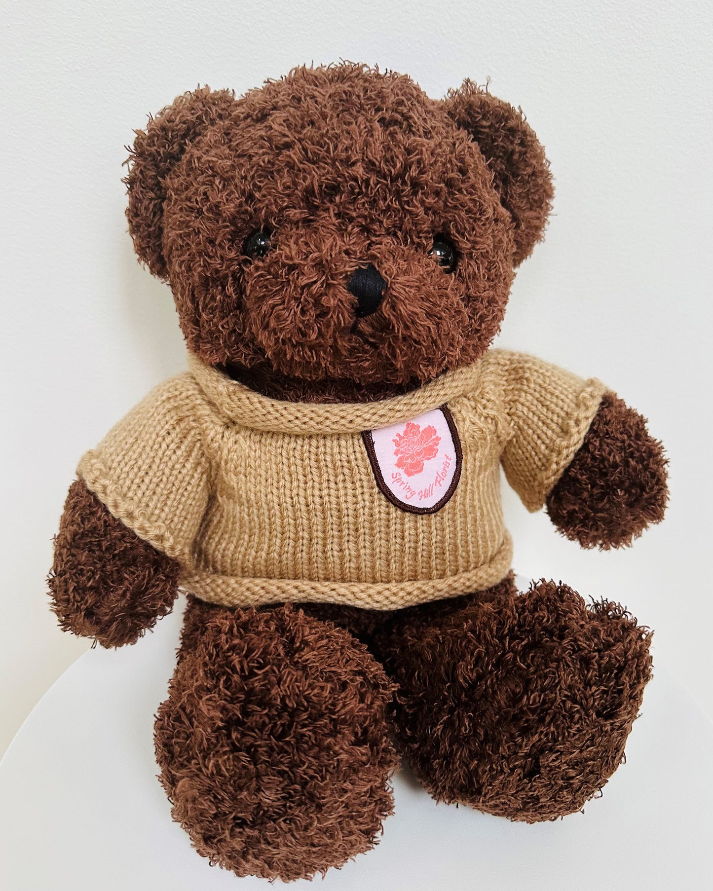 Soft brown teddy bear with curly fur, wearing a beige knitted sweater and pink floral badge.