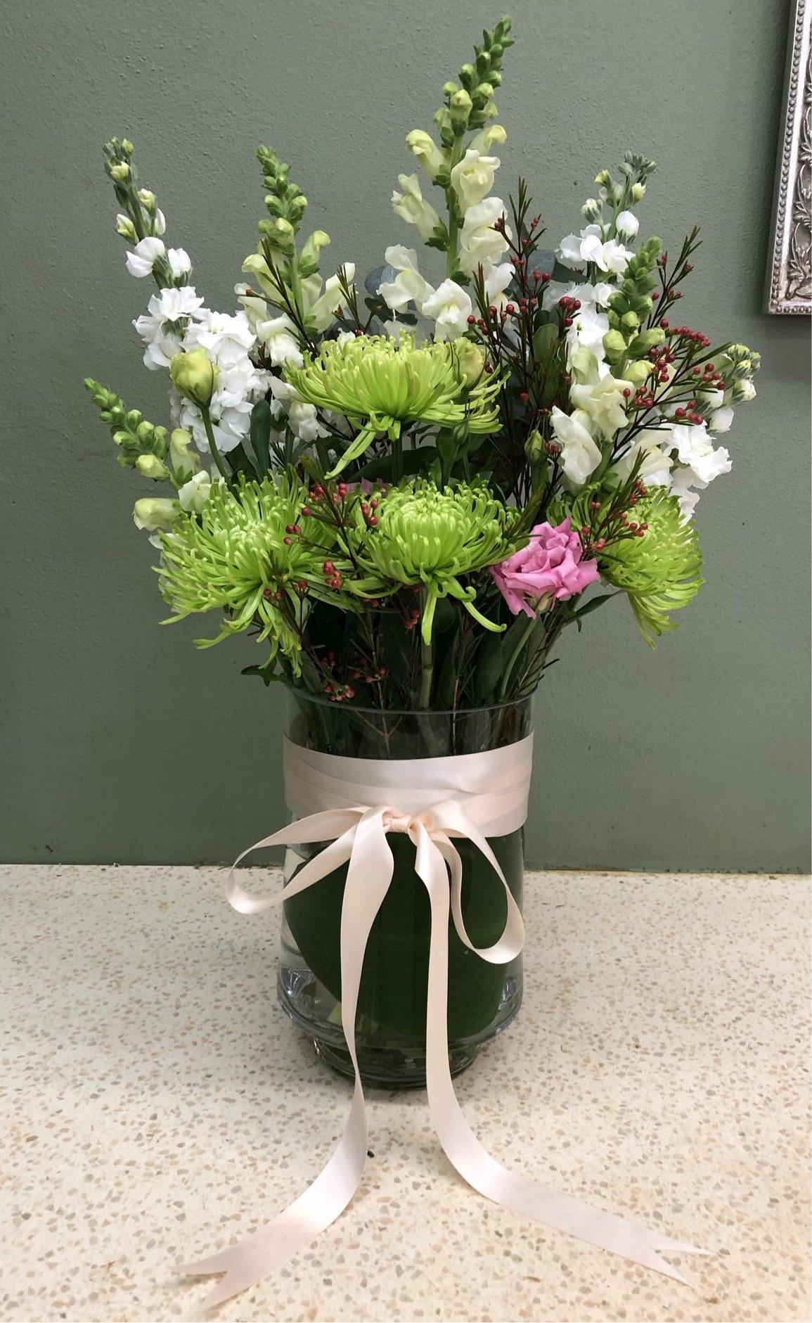 Fresh flower arrangement with green spider chrysanthemums, white snapdragons, and pink carnation in a glass vase with a satin ribbon bow, perfect for floral gifts or decor.