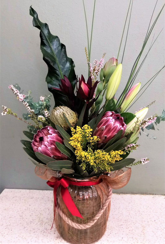 Native flower arrangement with protea, lilies, and greenery in a jar tied with a red ribbon