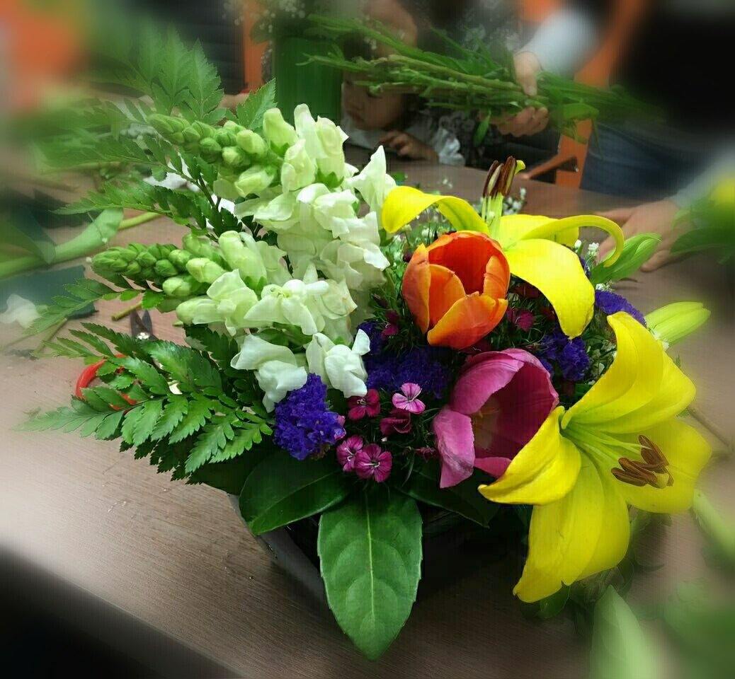 Bright mixed flower arrangement with yellow lilies, orange tulips, white snapdragons, ferns on a table.