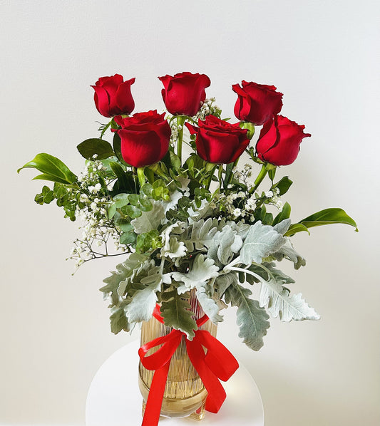 Six red roses in a clear vase with greenery, baby's breath, and a red ribbon