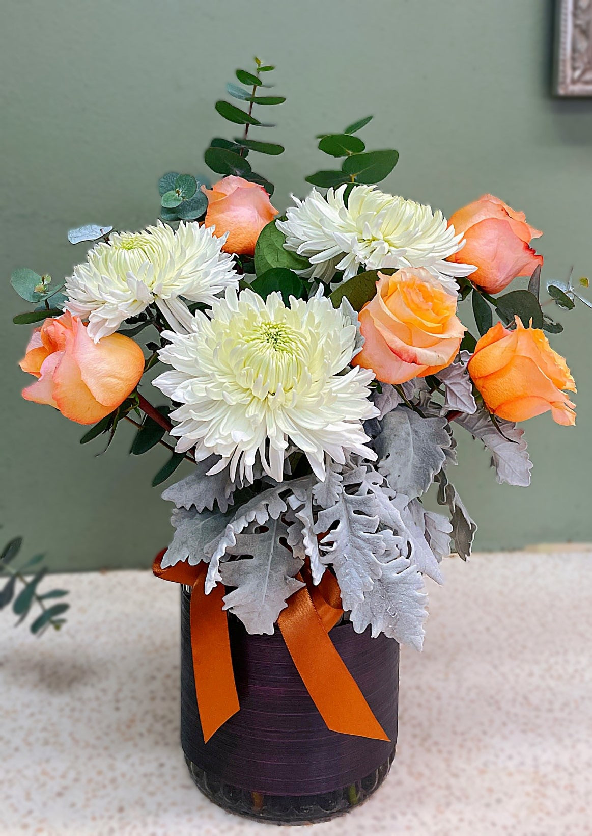 Peach roses and white chrysanthemums in a vase with greenery and silver leaves, orange ribbon