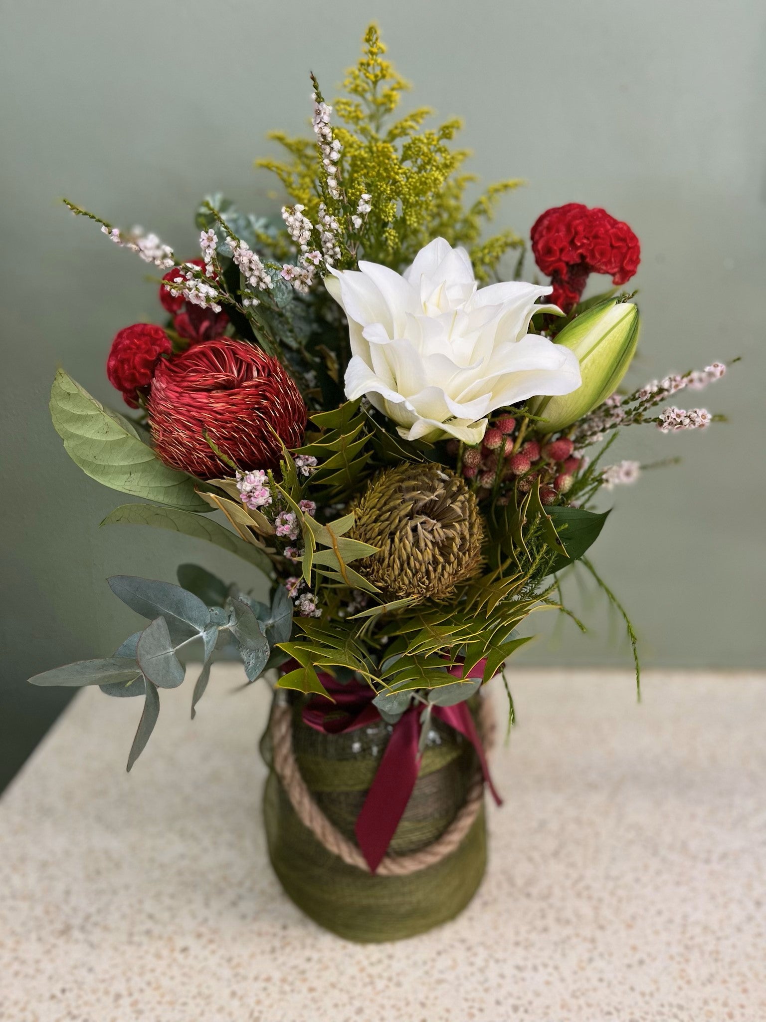 Native flower arrangement with white lily, red banksia, greenery in a rustic vase with rope accent.