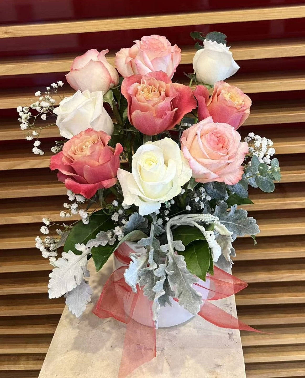 Pink and white rose bouquet with greenery and baby's breath in a white vase with a pink ribbon.
