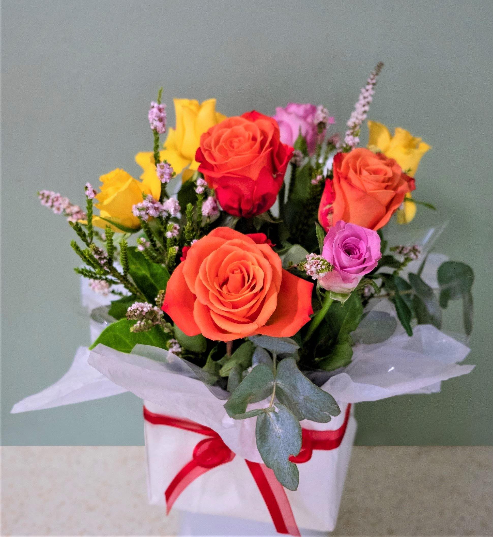 Gift box bouquet with orange, yellow, and pink roses, greenery, and red ribbon