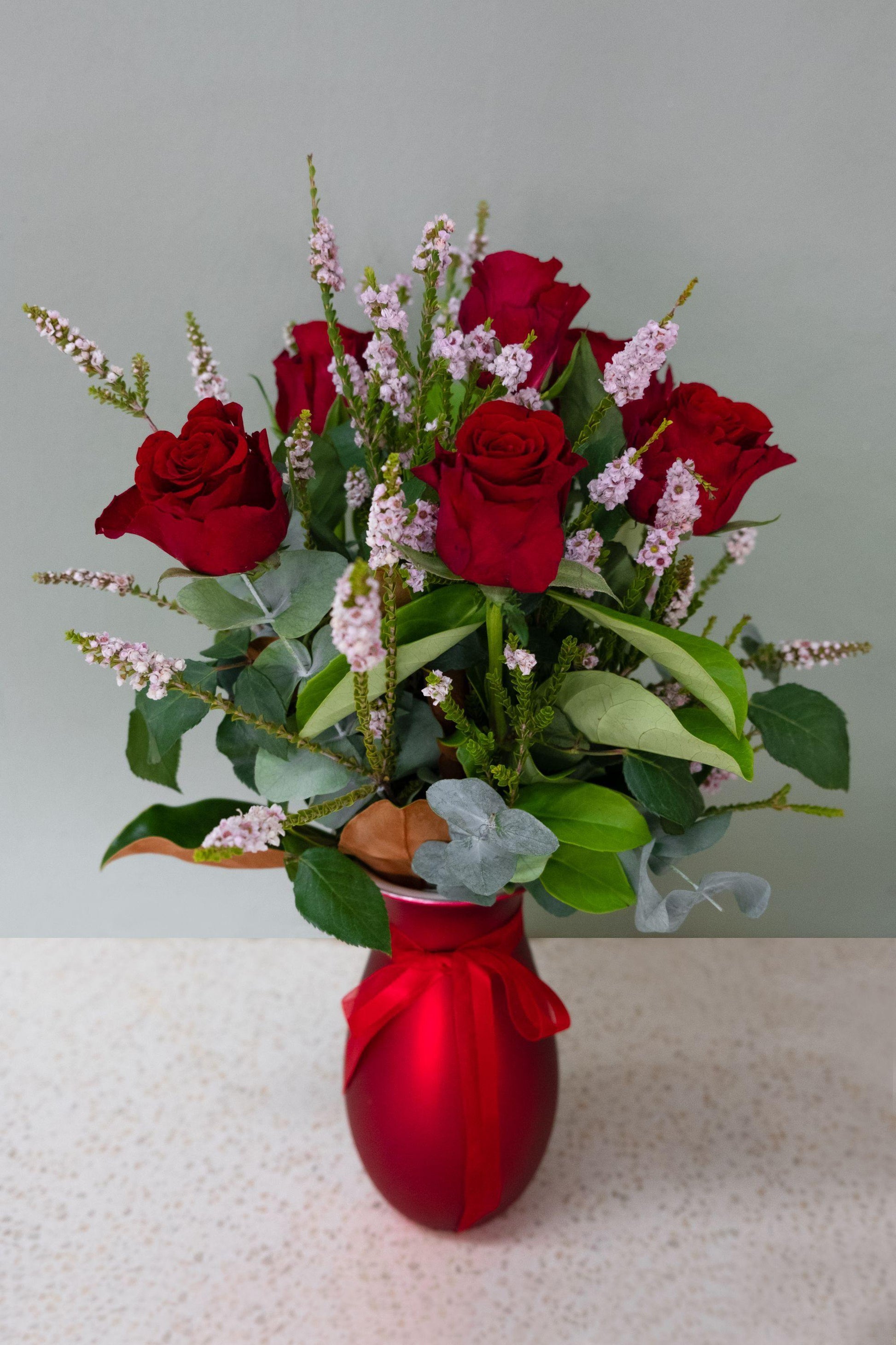 Red roses and pink flowers arranged in a red vase with greenery on a light table.