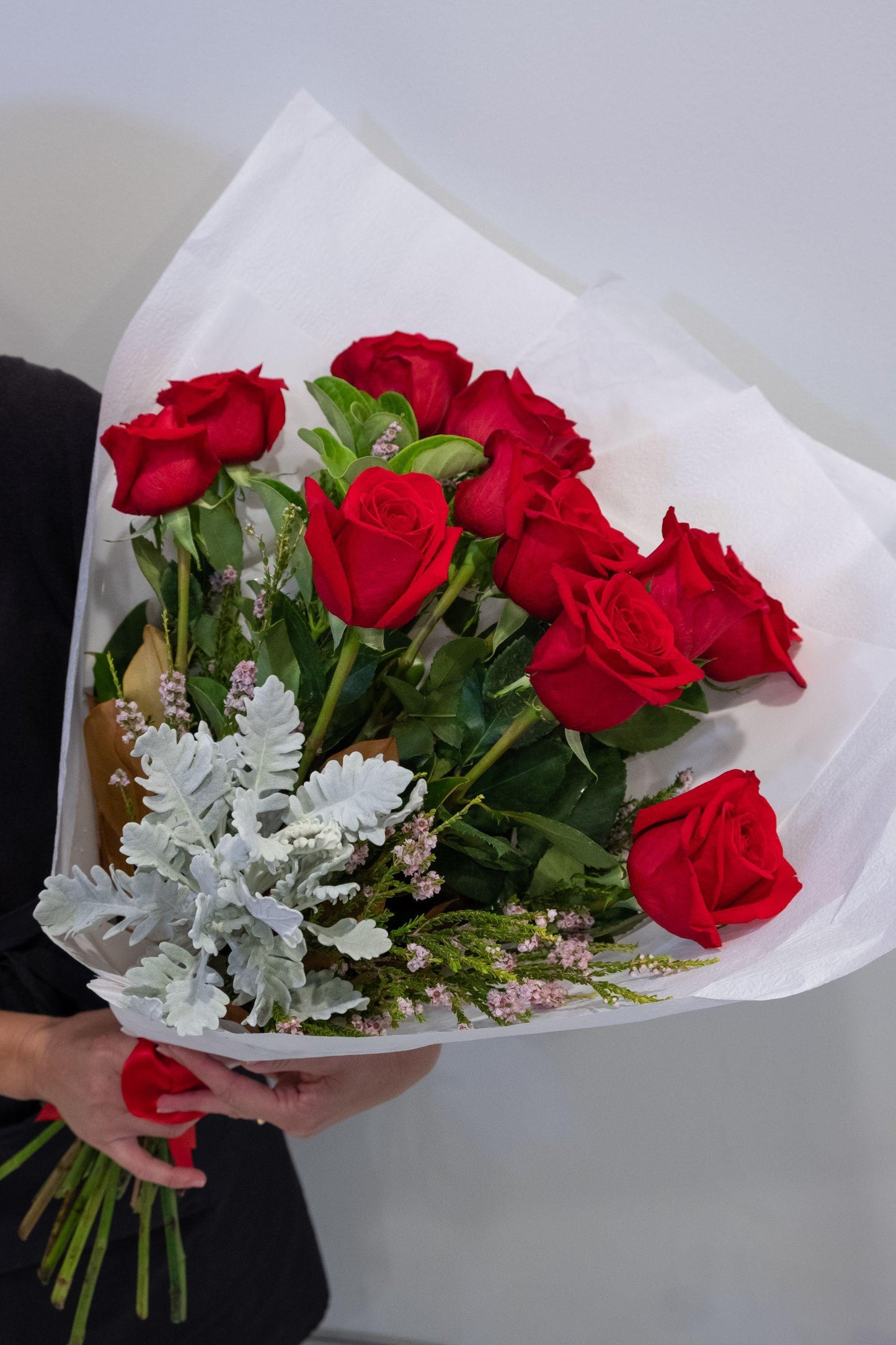 Hand holding a bouquet of fresh red roses with white and green foliage, wrapped in white paper.