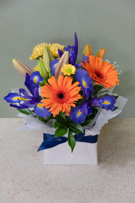 Bright mixed flower arrangement with orange gerberas, blue iris, lilies, and greenery in a gift box