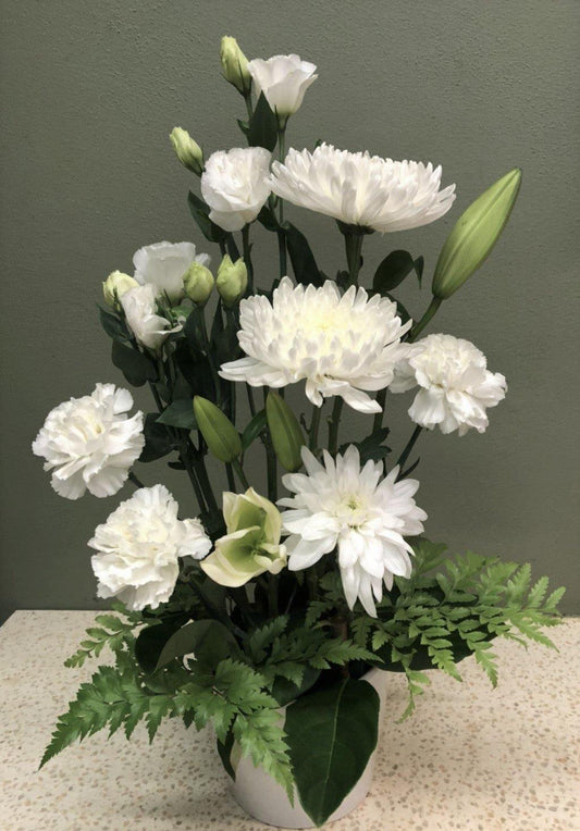 Elegant white flower arrangement with chrysanthemums, lilies, and lush green foliage in a white vase