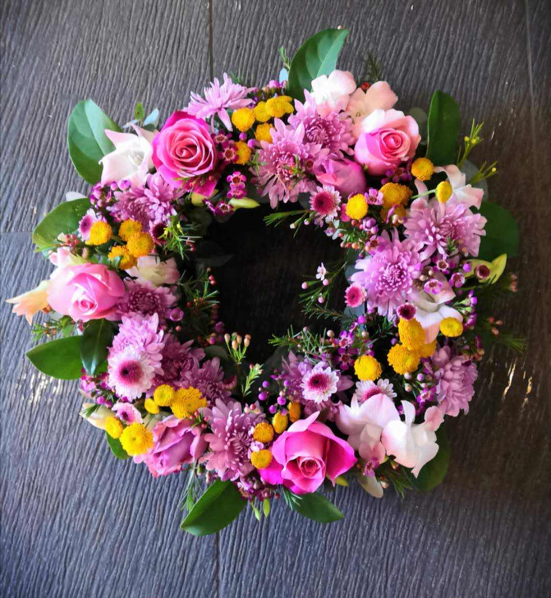 Colorful floral wreath with pink roses, yellow billy buttons, chrysanthemums and lush greenery on a dark textured surface.
